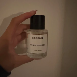 ESSNCE Summer Breeze parfym - Fräsch parfym från ESSNCE med namnet Summer Breeze. Flaskan är rund och genomskinlig med svart lock och vit etikett med stilren text. Innehållet är ljust och flaskan är i glas. Perfekt för dig som gillar lätta och somriga dofter.