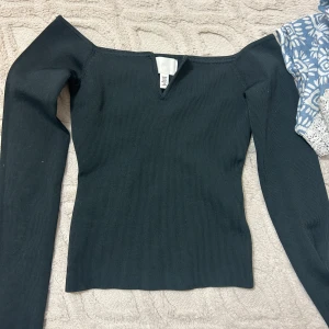 Svart ribbad offshoulder topp H&M - Svart ribbad topp från H&M med offshoulder-modell och lång ärm. Toppen har en snygg v-ringning framtill och sitter tight mot kroppen. Perfekt för dig som gillar stilrena och trendiga plagg.