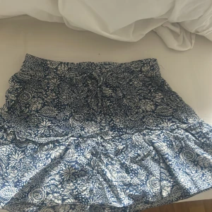 Blommig blåvit kortkjol med volang - Supersöt kortkjol i blått och vitt med blommigt mönster och volangkant. Kjolen har en bekväm midja med snörning och är gjord i ett lätt, mjukt bomullstyg som passar perfekt till varma dagar. Snygg att matcha med en enkel topp eller hoodie. Kjolen har inneshorts