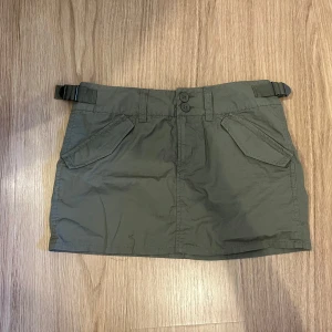 Grön kortkjol med fickor och bälte - Snygg militärgrön kortkjol med två stora fickor fram och två bak. Kjolen har dubbla knappar i midjan och justerbara bälten på sidorna för extra cool passform. Knapp använd och den är från hm.