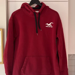 Röd hoodie från Hollister med tryck - Säljer en röd hoodie från Hollister med vit logga på bröstet och stort tryck på ryggen. Tröjan har huva med svarta snören, magficka och långa ärmar. Materialet är mjukt och känns fleece-aktigt, perfekt för kyliga dagar. Snygg och enkel att matcha med jeans eller joggers.