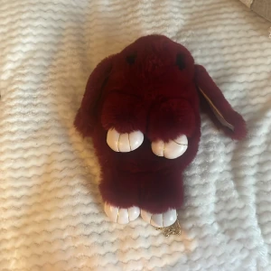 Vinröd kaninformad axelväska med kedja - Supergullig axelväska i form av en kanin, vinröd och fluffig med vita tassar och guldfärgad kedja. Väskan har dragkedja upptill och är tillverkad i mjuk syntetpäls. Perfekt för dig som vill sticka ut med en unik accessoar!