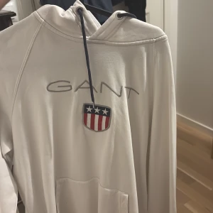 Vit hoodie från GANT med logga - Säljer en vit hoodie från GANT med broderad logga och emblem i rött, blått och vitt på bröstet. Tröjan har huva med mörkblå snörning och en stor magficka framtill. Perfekt för chill dagar och snygg streetstyle.