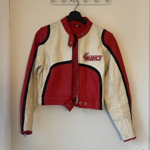 Röd och vit motorcykel acka Giudici - Säljer en riktigt snygg röd och vit motorcykeljacka i skinn från Giudici, tillverkad i Italien. Jackan har coola svarta detaljer, broderad logga på bröstet och dragkedja framtill. Den har långärmade ärmar med dragkedjor vid ärmsluten och en sportig, retro vibe.