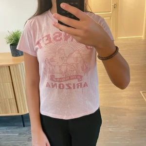 Ljusrosa Arizona t-shirt från H&M Divided - Snygg ljusrosa t-shirt från H&M Divided i storlek XXS. Tryck framtill med texten 'SUNSET IN THE DESERT OF ARIZONA' och motiv av berg och sol. T-shirten har rund hals, korta ärmar och är gjord i mjuk bomull. 