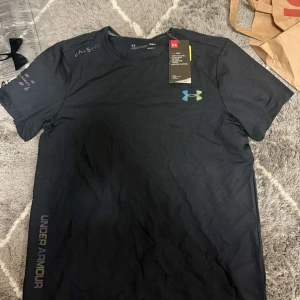 Svart t-shirt från Under Armour - Svart t-shirt från Under Armour med färgglad logga på bröstet och tryckt text på ärmen. Tillverkad i ett lätt och svalt syntetmaterial som passar perfekt för träning. Klassisk passform med korta ärmar och rund hals. Snygg och sportig vibe!