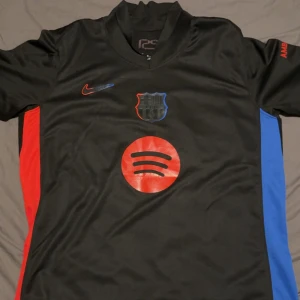 FC Barcelona Lamine Yamal tröja svart - Snygg svart FC Barcelona fotbollströja från Nike med Lamine Yamal och nummer 19 på ryggen. Tröjan har röda och blå detaljer på sidorna, Spotify-logga på bröstet och klubbmärke framtill. Tillverkad i lätt och ventilerande material, perfekt för match eller träning.