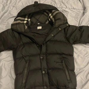 Svart pufferjacka från Burberry - Svart pufferjacka från Burberry med avtagbara ärmar så du kan bära den som väst. Klassisk rutig insida på huvan, dragkedja och knappar framtill samt flera fickor med dragkedja. Perfekt för kalla dagar och riktigt snygg streetstil,10/10 skick liten i storlek så kan passa XS.