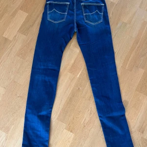 Jacob Cohën blå skinny jeans - Snygga blå jeans från Jacob Cohën med kontrastsydda sömmar och klassisk femficksdesign. Modellen är skinny med smal passform och raka ben. Jeansen har exklusiva detaljer som märkets logga på bakfickan och en unik knapp framtill. Perfekta för dig som gillar stilrena och lyxiga jeans.