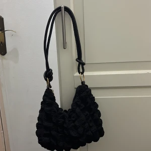 Svart quiltad axelväska med knutdetalj - Säljer en svart axelväska med quiltad yta och coola knutdetaljer på axelremmen. Väskan har guldfärgade ringar och dragkedja upptill. Materialet känns mjukt och har en unik struktur som sticker ut. Perfekt storlek för mobilen, plånboken och lite till.