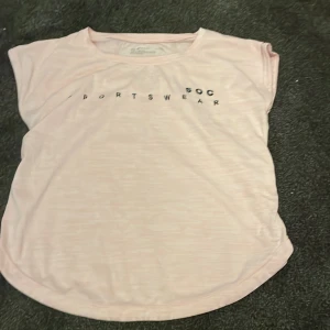 Ljusrosa t-shirt från SOC - Ljusrosa t-shirt från SOC med trycket 'SPORTSWEAR' framtill. Modellen är kortärmad och har en rundad nederkant. Materialet är mjukt och lätt polyester, perfekt för träning eller chill. Snygg och enkel design som funkar till många stilar.