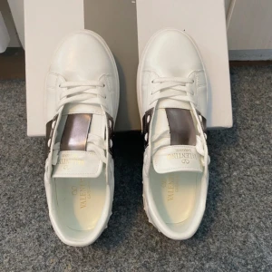 Valentino Garavani vita / silver  - Snygga vita sneakers från Valentino Garavani med bred metallic-silver rem över vristen och nitar på sidorna. Skorna har snörning, platt sula och är tillverkade i skinn.  ( använda bara 1 gång)