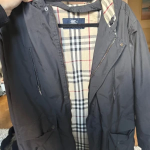 Svart vindjacka från Burberry - Snygg svart vindjacka från Burberry med klassiskt rutigt foder i beige, svart och rött. Jackan har dragkedja, fickor med lock och en stilren look. Perfekt för dig som vill ha en ikonisk jacka med lyxig känsla och street vibe.