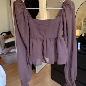 Brun blus från Abercrombie & Fitch - Säljer en brun blus från Abercrombie & Fitch med långa ärmar och volangdetaljer. Blusen har en luftig passform och är perfekt för en stilren look. Den har en fyrkantig halsringning och elastiska ärmslut.
