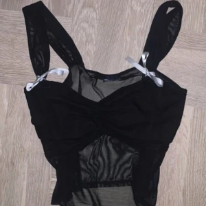 Svart mesh korsettopp - Snygg svart korsettopp i mesh med knytband på axlarna. Perfekt för en kväll ute eller när du vill känna dig extra speciell. Toppen har en åtsittande passform och en stilren design.