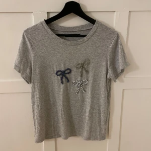 Grå t-shirt med pärlrosetter - Säljer en grå t-shirt från BikBok med tre dekorativa pärlrosetter i olika färger på framsidan. T-shirten har en rund hals och korta ärmar. 
