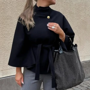 Svart kappa med knytband -  Helt ny med lappen på , passar ej i den !!!Elegant svart kappa med hög krage och knytband i midjan. Den har en knappdetalj vid halsen och vida ärmar som ger en stilren look. Perfekt för att höja din outfit till nästa nivå.