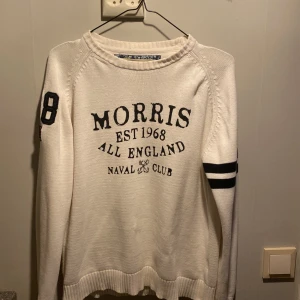 Vit tröja från Morris - Säljer en vit tröja från Morris med tryck 'Morris Est 1968 All England Naval Club' på framsidan. Tröjan har långa ärmar med svarta ränder och siffran 68 på ärmen. Perfekt för en stilren och avslappnad look.