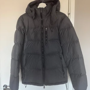 Grå dunjacka från Polo Ralph Lauren - Säljer en snygg grå dunjacka från Polo Ralph Lauren. Jackan har en dragkedja framtill och flera praktiska fickor. Perfekt för kyliga dagar med sin varma och bekväma design. Den har en diskret logga på bröstet.