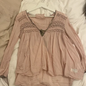 Rosa blus från Odd Molly - Säljer en söt rosa blus från Odd Molly med fina broderade detaljer och smock vid hals och ärmslut. Blusen har långa ärmar och en avslappnad passform. Perfekt för en bohemisk stil! 🌸