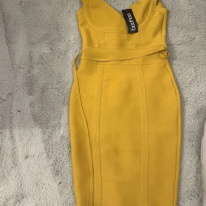 Gul klänning från Boohoo - Snygg gul klänning från Boohoo med en figurnära passform. Klänningen har breda axelband och en stilren design som framhäver figuren. Perfekt för en somrig look!
