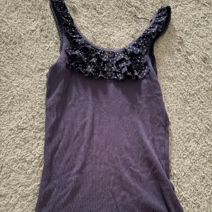 Lila linne från Atmosphere - Säljer ett lila linne från Atmosphere med volangdetaljer och prickigt mönster vid halsringningen. Linnet har en söt design med knappar framtill. Perfekt för en avslappnad stil.