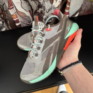 Reebok träningsskor  - Säljer ett par grå sneakers från Reebok med turkosa detaljer. Skorna har snörning och en sportig design, perfekt för träning. De har en slitstark sula och är gjorda i syntetmaterial.