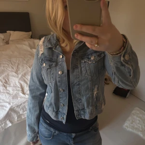 Blå croppad jeansjacka - Säljer en snygg blå croppad jeansjacka med slitna detaljer och knappar framtill. Jackan har en klassisk krage och långa ärmar. 