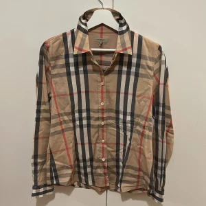 Rutig skjorta från Burberry - Säljer en klassisk rutig skjorta från Burberry i beige, svart och röd. Skjortan har långa ärmar och knappar framtill. Perfekt för en stilren look.