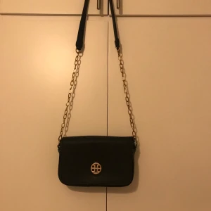 Svart axelväska med kedjedetalj - Snygg svart axelväska med guldfärgad kedja och ett elegant emblem på framsidan. Perfekt för att ge din outfit en stilfull touch. Väskan har en praktisk storlek och en justerbar rem för bekväm användning.