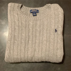 Grå stickad tröja från Polo Ralph Lauren - Säljer en stilren grå stickad tröja från Polo Ralph Lauren med kabelstickat mönster. Tröjan har långa ärmar och en rund halsringning. Perfekt för kyligare dagar. 🐎