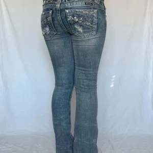 Blå jeans med broderade detaljer från Miss Me - Snygga blå jeans från Miss Me med broderade detaljer och gnistrande dekorationer på bakfickorna. De har en bootcut-stil och låg midja, vilket ger en trendig look. Perfekta för att lägga till lite extra stil i din garderob. Innerbenslängd 76cm och midjemått rakt över 35cm 💋