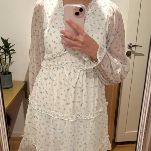 Vit blommig klänning med volangdetaljer - Vit klänning med blå blommor och volangdetaljer. Den har långa, transparenta ärmar och en omlott-design framtill, går ned precis ovanför knäna. Endast använd en gång 