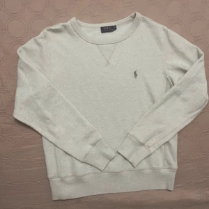Beige tröja från Ralph Lauren - Säljer en stilren beige tröja från Ralph Lauren med rund hals och långa ärmar. Tröjan har en liten broderad logga på bröstet och ribbade muddar vid ärmslut och nederkant. Perfekt för en avslappnad look.