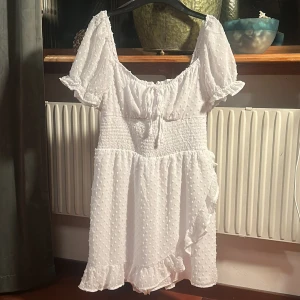 Vit kortklänning - Säljer en söt oanvänd vit kortklänning med puffiga kortärmar och smockdetaljer i midjan. Klänningen har ett diskret prickigt mönster och en söt rosett framtill. Klänningen är tunt sydd med shorts intill för att förebygga att den åker upp. Perfekt för sommarens varma dagar!