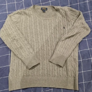 Grå stickad tröja från Polo Ralph Lauren - Säljer en stilren grå stickad tröja från Polo Ralph Lauren med klassiskt flätmönster. Tröjan har långa ärmar och en broderad logotyp på bröstet. Perfekt för kyliga dagar! Jag är 180 och den passar bra. Kan passa folk som är en del längre och en del kortare!🤩