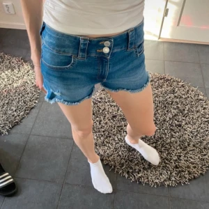 Blå jeansshorts  - Snygga blå jeansshorts med fransiga kanter och två knappar framtill. Perfekta för en avslappnad stil. De har en klassisk femficksdesign och är bekväma att bära. Storlek 170 men sitter som xs