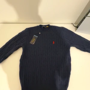 Mörkblå stickad tröja från Polo Ralph Lauren - Säljer en snygg mörkblå stickad tröja från Polo Ralph Lauren med kabelstickat mönster. Tröjan har en rund halsringning och ett broderat rött logotypmärke på bröstet. Perfekt för en stilren look.