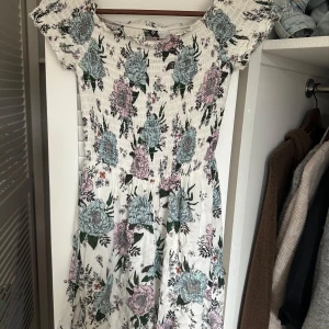 Blommig klänning från Divided - Säljer en söt blommig klänning från Divided. Den har en smockad överdel och korta ärmar. Klänningen är vit med blommor i blått och lila. Perfekt för sommarens alla tillfällen! 🌸