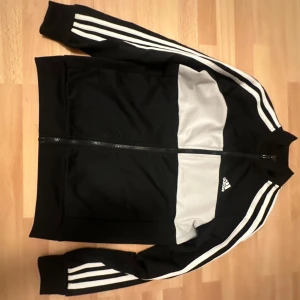 Svarta träningsbyxor från Adidas - Snygga svarta träningsbyxor från Adidas med vita ränder längs sidorna. Byxorna har en smal passform och är perfekta för sport och träning. De är tillverkade i ett bekvämt syntetmaterial och har en elastisk midja för bästa komfort.