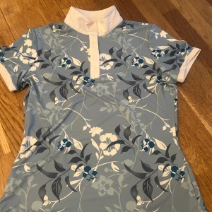 Blommig pikétröja från HKM - Säljer en stilren ljusblå pikétröja från HKM med ett vackert blommönster i vitt och mörkblått. Tröjan har korta ärmar och en klassisk krage med knappar. Perfekt för en avslappnad och elegant look.