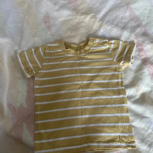 Gul och vit randig t-shirt från Primark - Säljer en söt gul och vit randig t-shirt från Primark för små barn. T-shirten har korta ärmar och är tillverkad i mjukt bomullsmaterial. Perfekt för varma dagar!