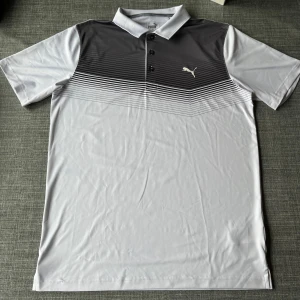 Grå och vit pikétröja från Puma - 📦Säljer en stilren grå och vit pikétröja från Puma. Tröjan har ett diskret randigt mönster upptill och en klassisk krage med knappar. Perfekt för en sportig look eller en avslappnad dag. (Storlek M, normal i storleken) Tröjan är i princip oanvänd och har inga defekter. Vid frågor, tveka inte att höra av er! ☺️