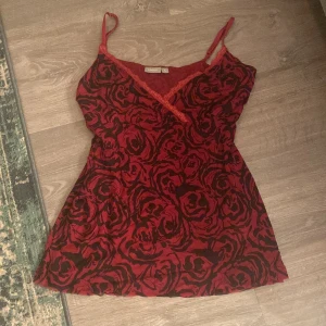 Röd blommig linne från Lindex - Säljer ett rött linne från Change med blommigt mönster och spetsdetaljer. Linnet har smala axelband och en v-ringad design. 