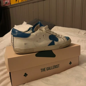  The Gallerist storlek 44 - Snygga vita sneakers med blå detaljer från The Gallerist. Skorna har en klassisk snörning och en stilren design med blå detaljer på häl och tunga. I storlek 44
