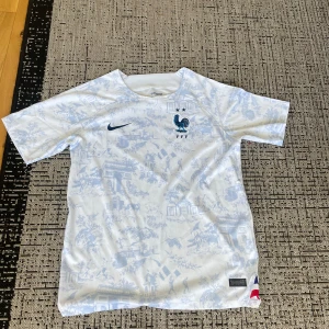 Frankrike Nike fotbollströja vit/blå - Säljer en officiell Frankrike fotbollströja från Nike i vit med ljusblå mönster. Tröjan har korta ärmar, rund hals och detaljer med franska flaggans färger vid nederkanten. Materialet är lätt och andas, perfekt för match eller träning.