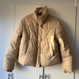 Beige dunjacka från NLY Trend - Snygg beige pufferjacka från NLY Trend i storlek 38. Jackan har hög krage, två fickor framtill och quiltad design. Perfekt för kalla dagar med sin oversized look och mjuka material. Enkel att matcha med allt i garderoben.