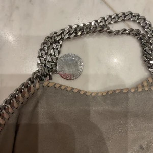 Grå axelväska från Stella McCartney - Säljer en grå axelväska från Stella McCartney med grova silverfärgade kedjor runt kanterna och som axelrem. Väskan är tillverkad i syntetmaterial med ljusbeige detaljer längs sömmarna. Insidan har ett ljust foder med logotypmönster och en innerficka med dragkedja.