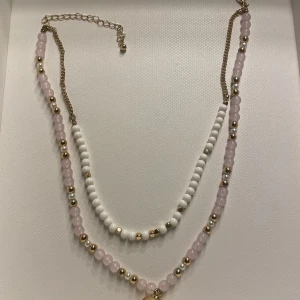 Dubbel halsband med svampberlock från H&M - Säljer ett dubbel halsband med pärlor i ljuslila, vitt och guld. Ena kedjan har vita pärlor och små guldfärgade detaljer, den andra har ljuslila pärlor och guldfärgade kulor samt en gullig svampberlock i guld. Perfekt för att ge din outfit en söt och lekfull touch. Använd ett fåtal gånger och har fått små slitningar på berlocken, men inget som syns tydligt