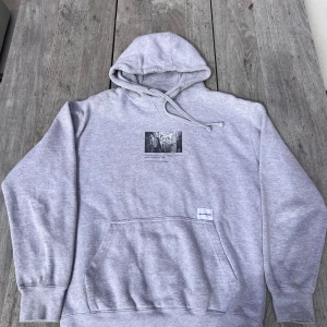 Grå hoodie MKI Miyuki Zoku med print - Säljer en grå hoodie från MKI Miyuki Zoku med coolt svart print på ryggen och ett foto med text på bröstet. Tröjan har huva med snörning, stor magficka och ribbade muddar. Perfekt för dig som gillar streetwear och vill ha en chill look.